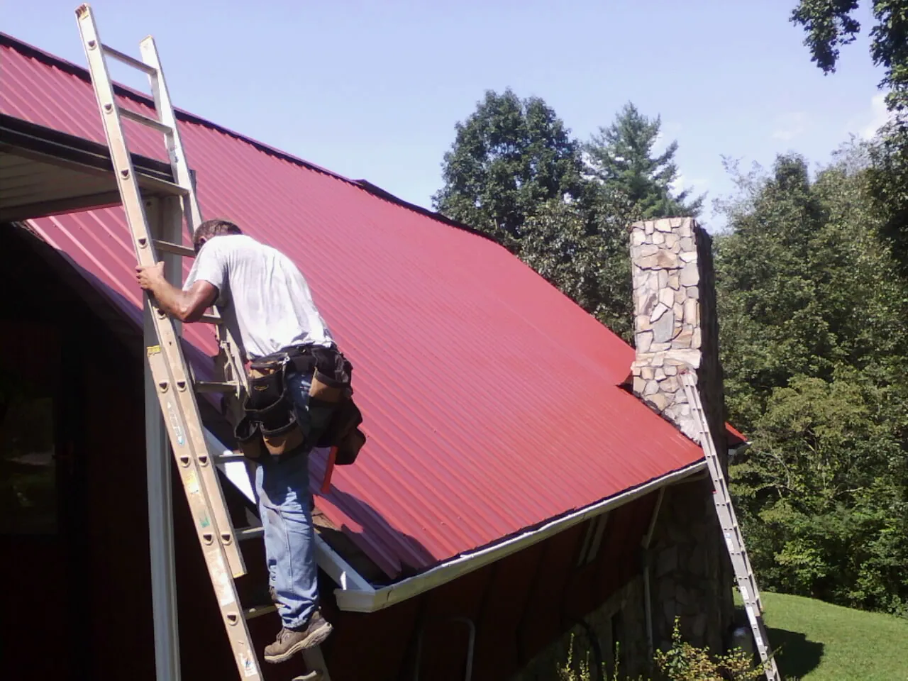 Roofer on a ladder beside a red metal roof and stone chimney — Soffit & Fascia Repair by Roofing Brothers in Marysvale